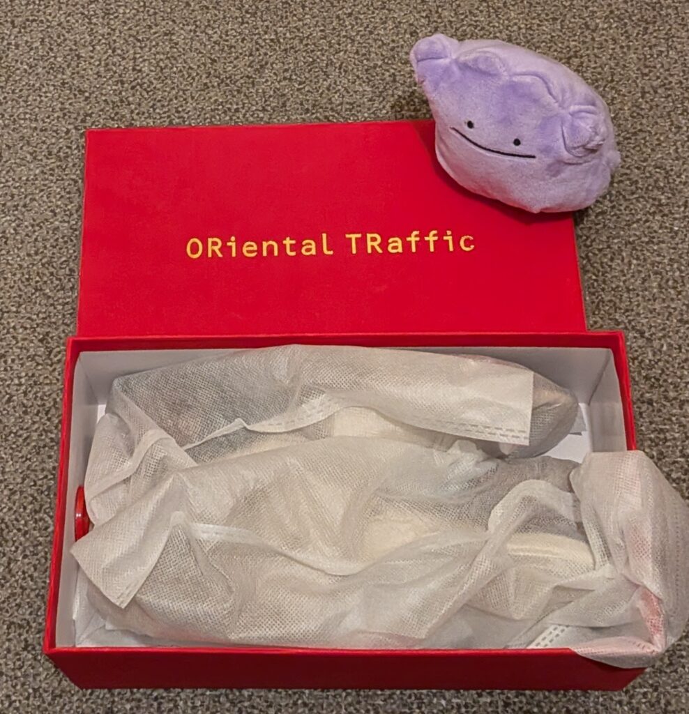 ダブルエーの株主優待で届いたORiental TRafficの赤い靴箱と、それを見守るメタモンのぬいぐるみ